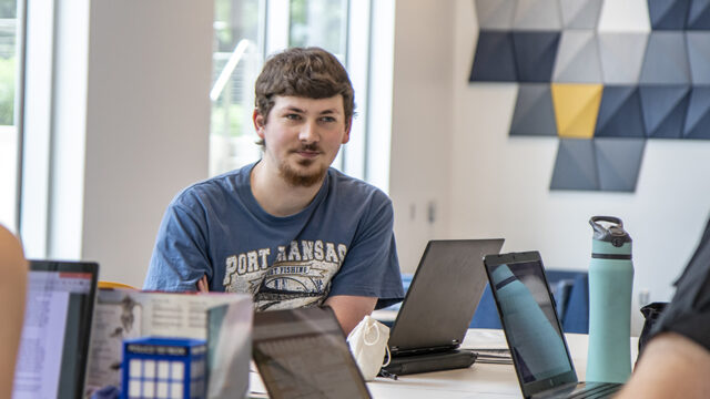 Student sitting at a table with several laptops on the table