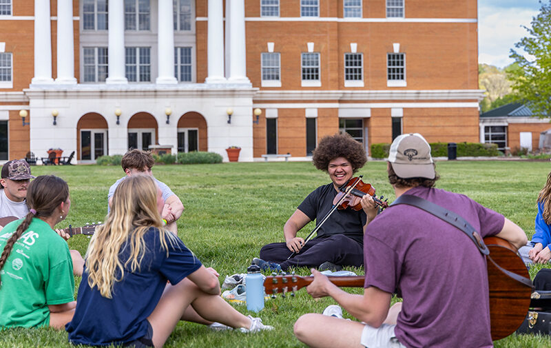 Group of students sitting a circle on the grass playing music together