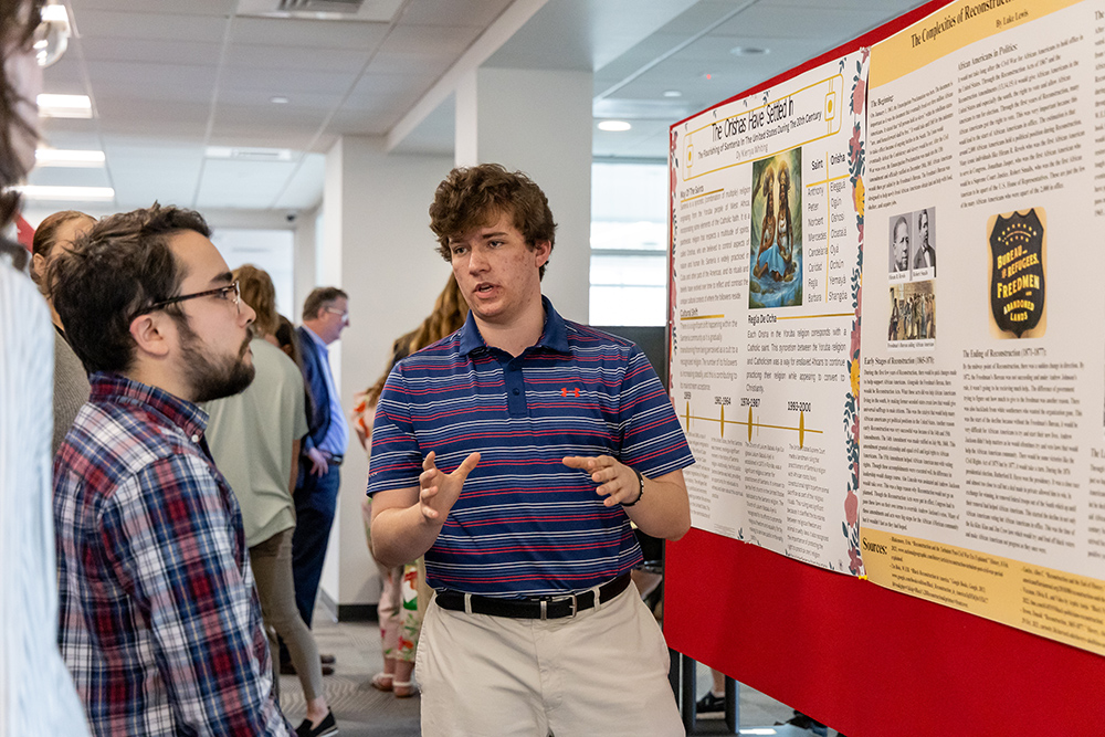 Student wearing striped polo talking to someone else in front of a poster presentation