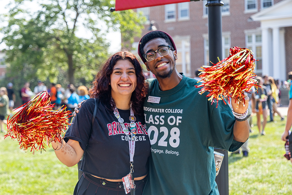 Two students standing next to each other smiling and waving gold and crimson pom-poms