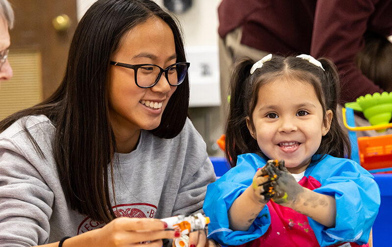 Female student smiling at young child who's hands are covered in black paint