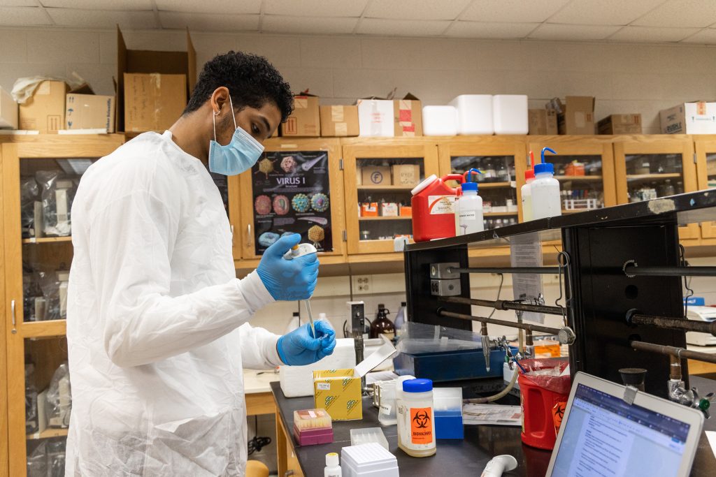 Student wearing white shirt with mask and gloves using equipment in biology lab