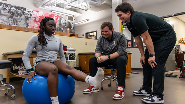 Two athletic trainers observing an athlete that is sitting on a large blue ball with one leg extended