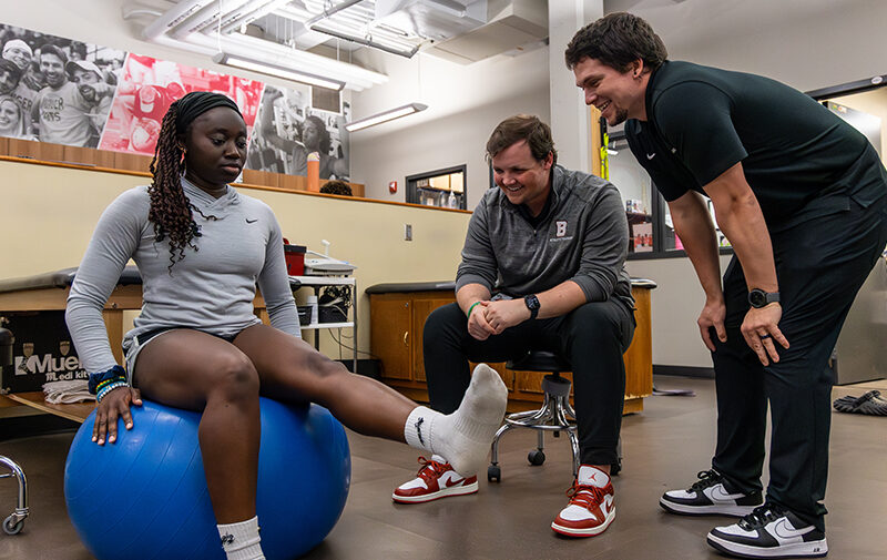 Two athletic trainers observing an athlete that is sitting on a large blue ball with one leg extended