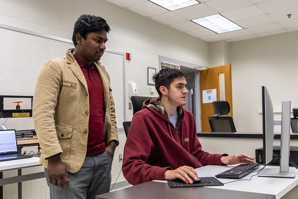 Professor standing behind engineering student that is sitting down looking at a computer screen