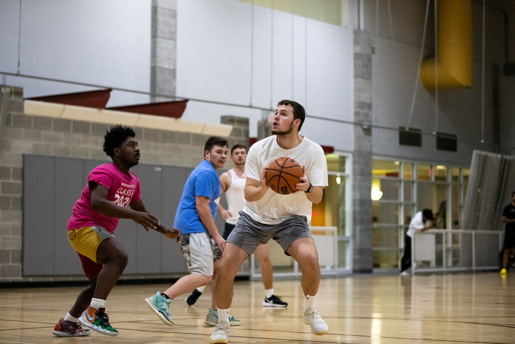 Students playing intramural basketball. One man is shooting the basketball wearing a white shirt.