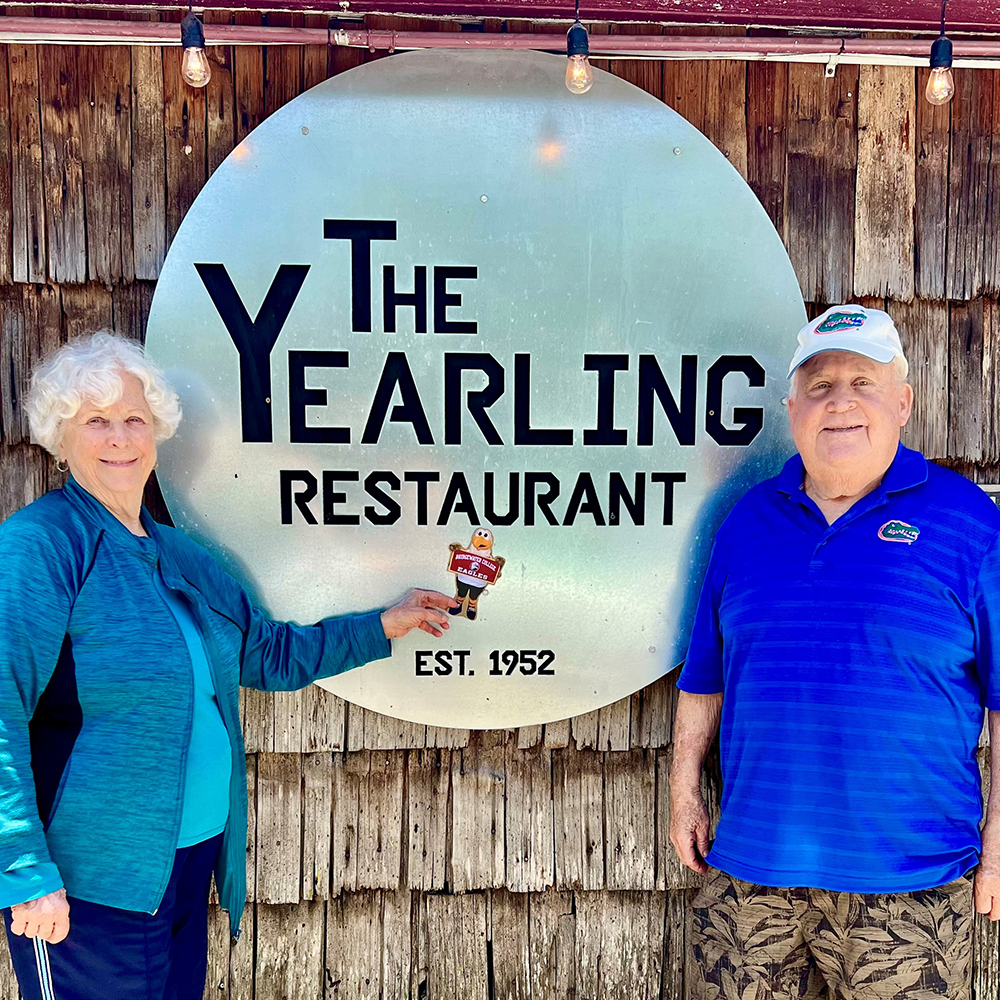 Woman holding Flat Ernie in front of metal sign that reads The Yearling Restaurant. Man in blue standing to the right of the sign. 