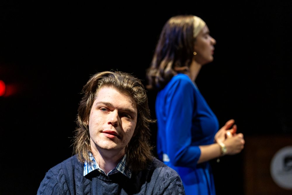 An man monologues while a woman paces behind him