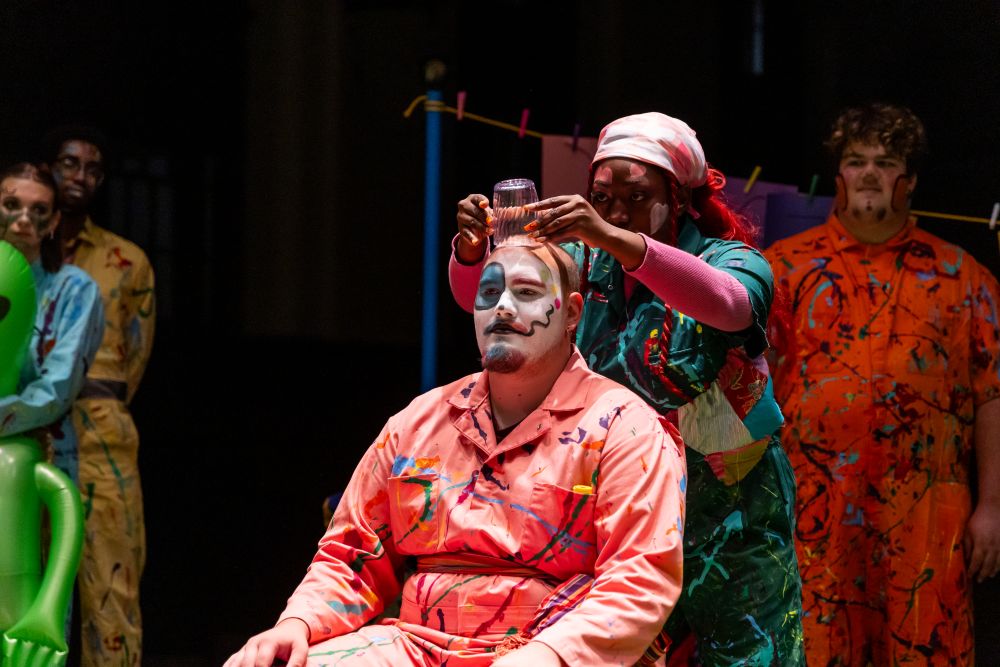 A woman sets a glass on top of a man's head. Both characters are painted in absurd color cobinations