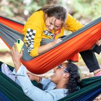 Two girls in stacked hammocks look at a phone and laugh
